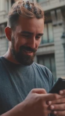 VERTICAL VIDEO: Young smiling man with beard uses smartphone on road background