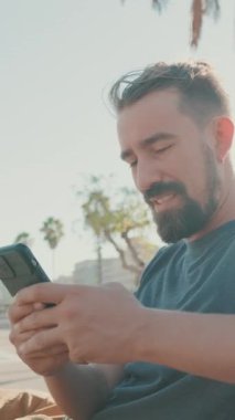 VERTICAL VIDEO: Young smiling man with beard sits on bench, uses cellphone