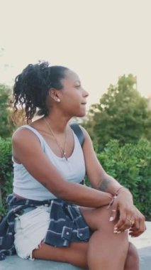 VERTICAL VIDEO: Young mixed race woman sitting on park bench, smiling, looking around