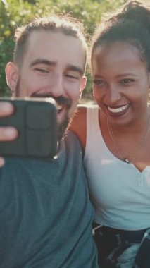 VERTICAL VIDEO: Happy smiling interracial couple taking selfie on phone while sitting on park bench. Backlight