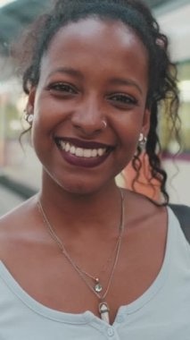 VERTICAL VIDEO: Close-up of smiling young mixed race woman standing at railway station on trains background