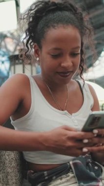 VERTICAL VIDEO: Young mixed race woman sitting at the railway station, using cellphone