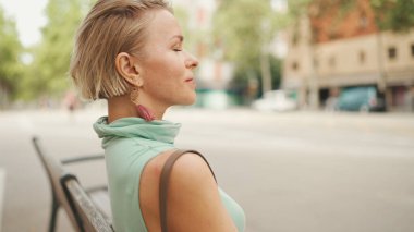 Beautiful woman with short blond hair in casual clothes sits on bench, smiles, enjoys warm summer day