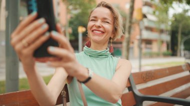 Beautiful happy smiling woman with short blond hair in casual wear sits on bench, makes video call on cellphone