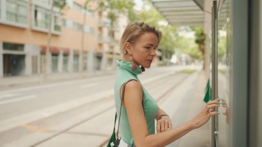 Beautiful woman with short blond hair in casual clothes stands at tram stop, buys ticket to travel on public transport