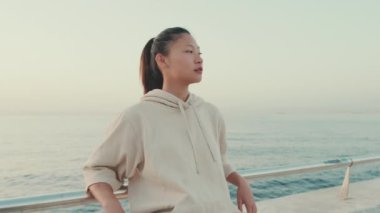 Asian girl wearing sportswear sits on the waterfront, enjoys the fresh air at morning time. Camera moving back