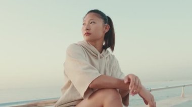 Asian girl wears sportswear sits on the promenade at morning time, on the background of the sea,turns her head and looks at the camera