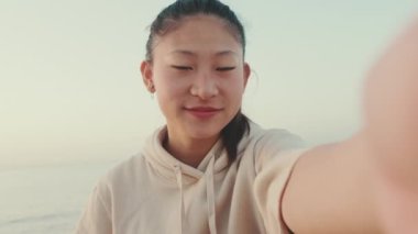 Smiling Asian girl wears sportswear sits on the promenade, takes selfie at morning time on the sea background