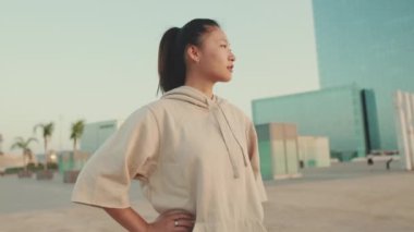 Asian girl in sportswear stands at morning time on modern buildings background