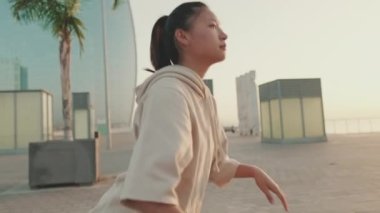 Asian girl in sportswear doing workout, squats, quadriceps exercises on modern buildings background at morning time