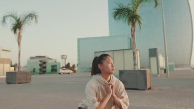 Asian girl in sportswear doing workout, squats, quadriceps exercises on modern buildings background at morning time
