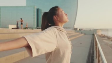 Asian girl in sportswear doing workout, breathing exercises, relaxing on modern buildings background at morning time
