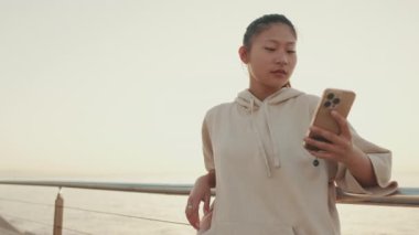 Asian girl in sportswear uses the phone while standing on the embankment in the morning