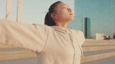 Asian girl in sportswear doing workout, breathing exercises, relaxing on modern buildings background at morning time