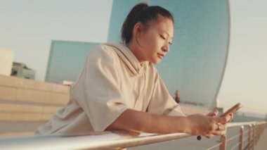 Smiling asian girl in sportswear uses phone while standing on waterfront on modern buildings background in morning light