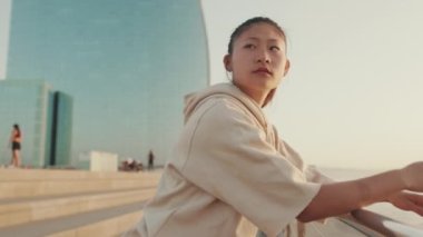 Asian girl in sportswear is resting in the training field, standing on modern buildings background at morning time
