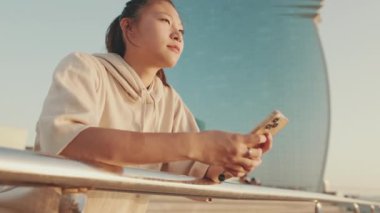 Asian girl in sportswear uses phone while standing on modern buildings background in morning light