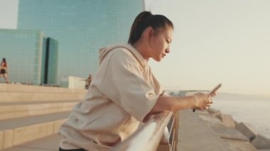 Smiling asian girl in sportswear uses phone while standing on waterfront on modern buildings background in morning light