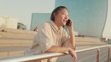 Smiling Asian girl in sportswear talking on the phone while standing on the embankment on modern buildings background in the morning light