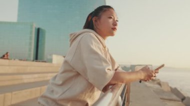 Asian girl in sportswear uses phone while standing on waterfront on modern buildings background in morning light