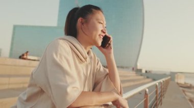 Smiling Asian girl in sportswear talking on the phone while standing on the embankment on modern buildings background in the morning light