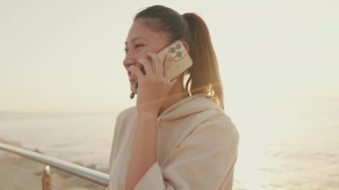 Close up, smiling Asian girl in sportswear talking on the phone while standing on the promenade on the sea background