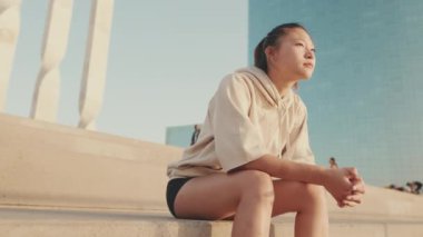 Asian girl in sportswear resting after workout sitting on steps on modern buildings background at sunrise