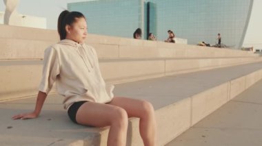 Asian girl in sportswear resting after workout sitting on steps on modern buildings background at sunrise