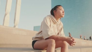 Asian girl in sportswear resting after workout sitting on steps on modern buildings background at sunrise
