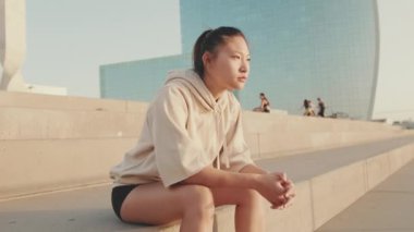 Asian girl in sportswear resting after workout while sitting on steps on modern buildings background at sunrise. Young woman turns her head and looks at the camera. Forward movement