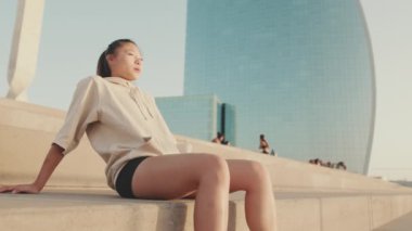 Asian girl in sportswear resting after workout sitting on steps on modern buildings background at sunrise