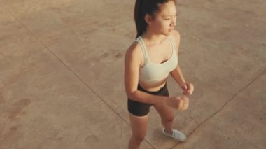 Asian girl in sports top does workout, squats, quadriceps exercises at morning time. View from above