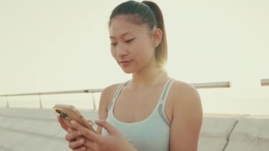 Close up, asian girl in sports top sits on the waterfront, uses smartphone. Backlight