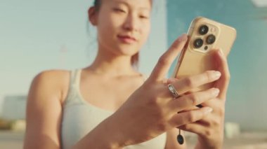 Close-up of smiling asian girl in sports top using cellphone while standing on waterfront on modern buildings background in morning light