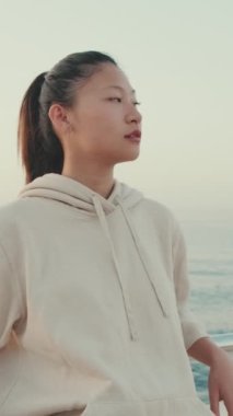 VERTICAL VIDEO: Asian girl wearing sportswear sits on the waterfront, enjoys the fresh air at morning time. Camera moving back