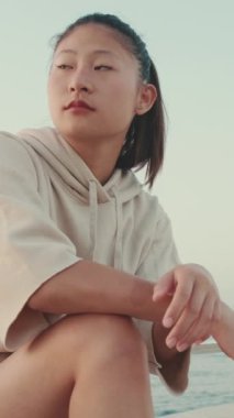 VERTICAL VIDEO: Asian girl wears sportswear sits on the promenade at morning time, on the background of the sea