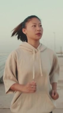 VERTICAL VIDEO: Asian young athletic woman running along the promenade. Sportswoman in sportswear exercising outdoors at morning time