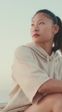VERTICAL VIDEO: Asian girl wears sportswear sits on the promenade at morning time, on the background of the sea,turns her head and looks at the camera