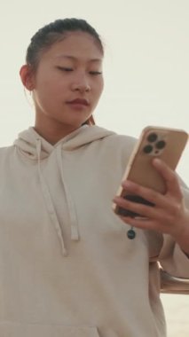 VERTICAL VIDEO: Asian girl in sportswear uses the phone while standing on the waterfront at morning time