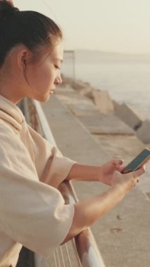 VERTICAL VIDEO: Smiling asian girl in sportswear uses phone while standing on waterfront on modern buildings background in morning light