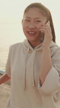 VERTICAL VIDEO: Smiling Asian girl in sportswear talking on the phone while standing on the embankment on modern buildings background in the morning light