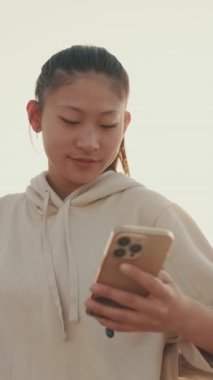VERTICAL VIDEO: Asian girl in sportswear uses the phone while standing on the waterfront at morning time