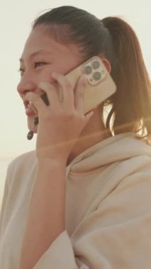 VERTICAL VIDEO: Smiling Asian girl in sportswear talking on the phone while standing on the embankment on modern buildings background in the morning light