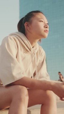 VERTICAL VIDEO: Asian girl in sportswear resting after workout sitting on steps on modern buildings background at sunrise