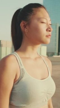 VERTICAL VIDEO: Asian girl in sports top stands on modern buildings background, turns her head and looks at the camera