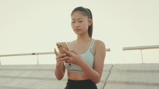 Asian Girl Sports Top Sits Waterfront Uses Smartphone — ストック動画