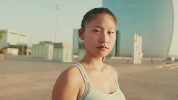 Asian Girl Sports Top Stands Modern Buildings Background Looks Camera — Video
