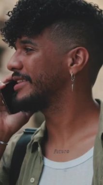 VERTICAL VIDEO: Portrait, close up of young happy man with beard dressed in an olive-colored shirt is talking on cellphone