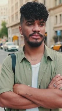 VERTICAL VIDEO: Young happy man with beard dressed in an olive-colored shirt is standing next to the road looking at the camera