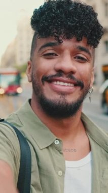 VERTICAL VIDEO: Young happy man with beard, dressed in an olive-colored shirt, stands next to the road, smiles and takes selfie